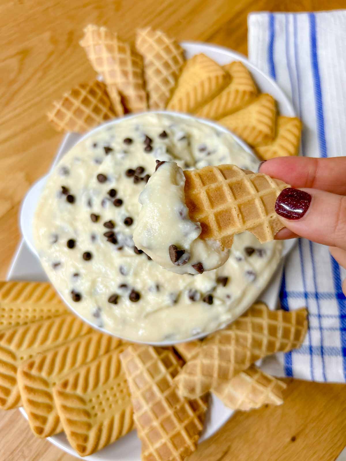 a white bowl is filled with cannoli dip and there are cookies around the bowl. a dipped cookie is held close to camera