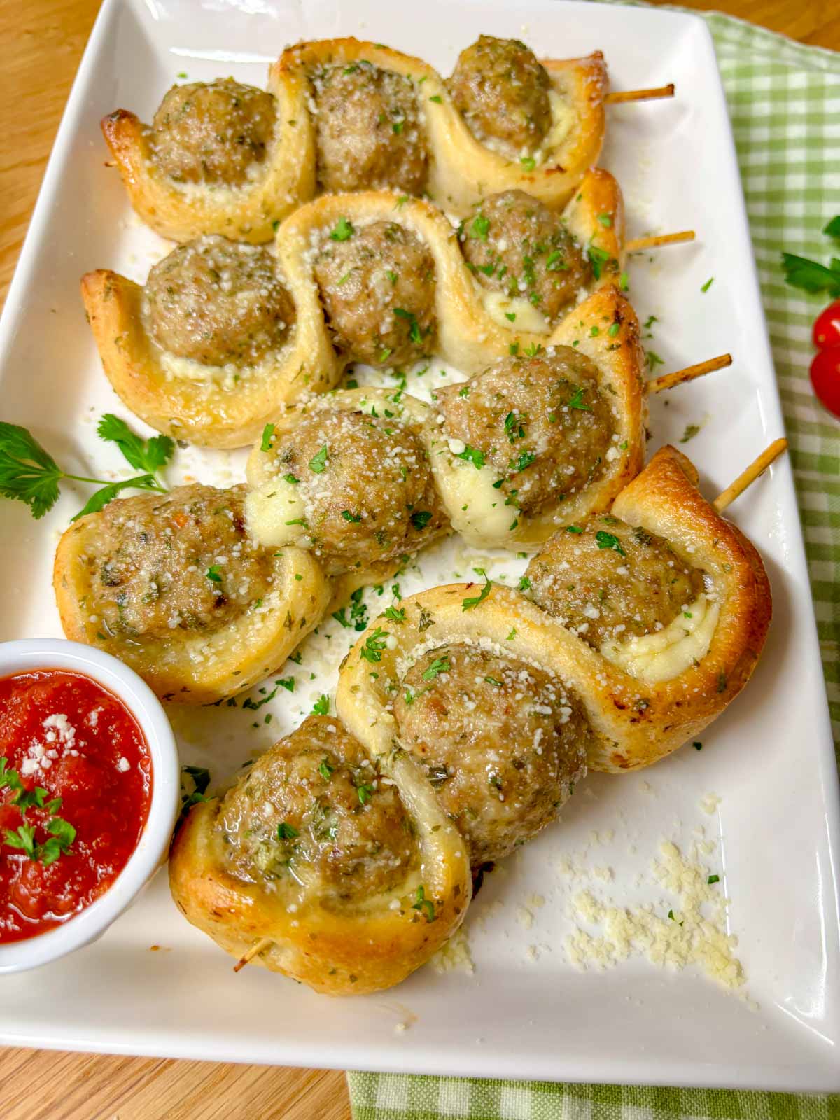 a white plate has chicken parmesan meatball subs on a stick with a side of marinara sauce to dip. garnished with parsley and parmesan cheese.