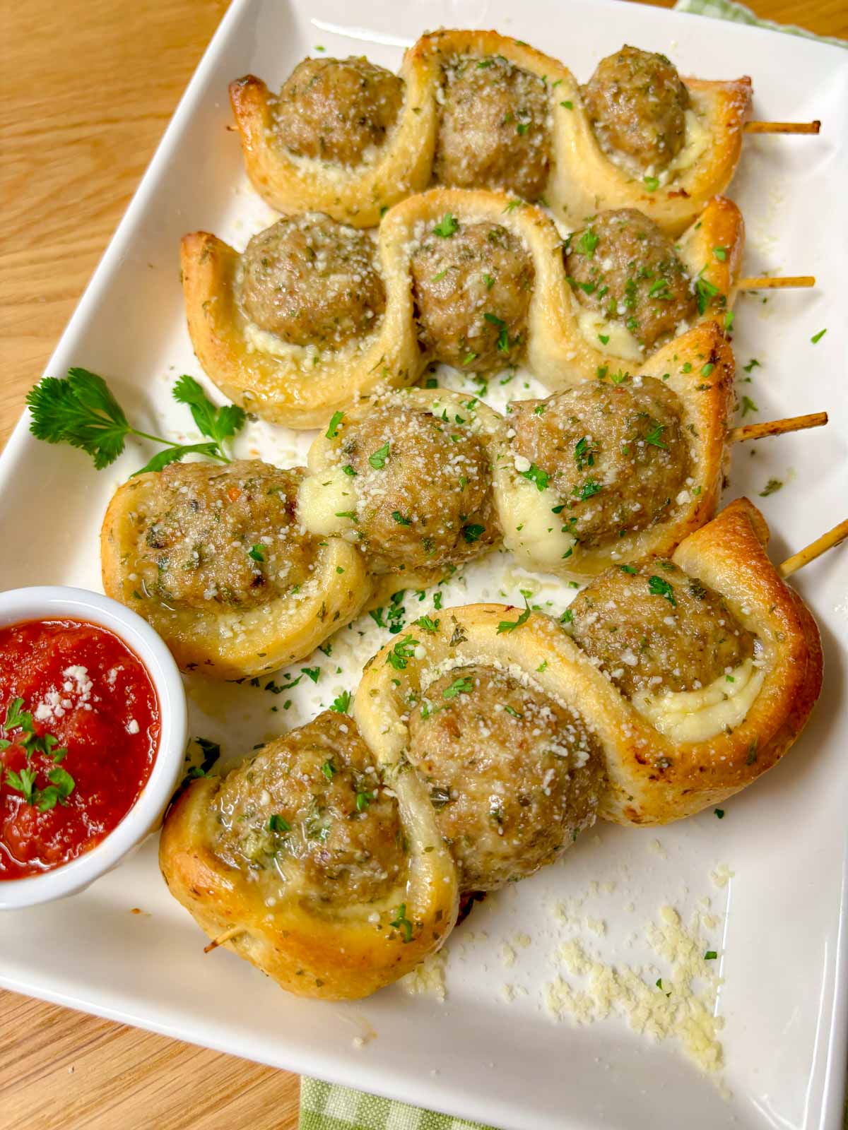 a white plate has chicken parmesan meatball subs on a stick with a side of marinara sauce to dip. garnished with parsley and parmesan cheese.