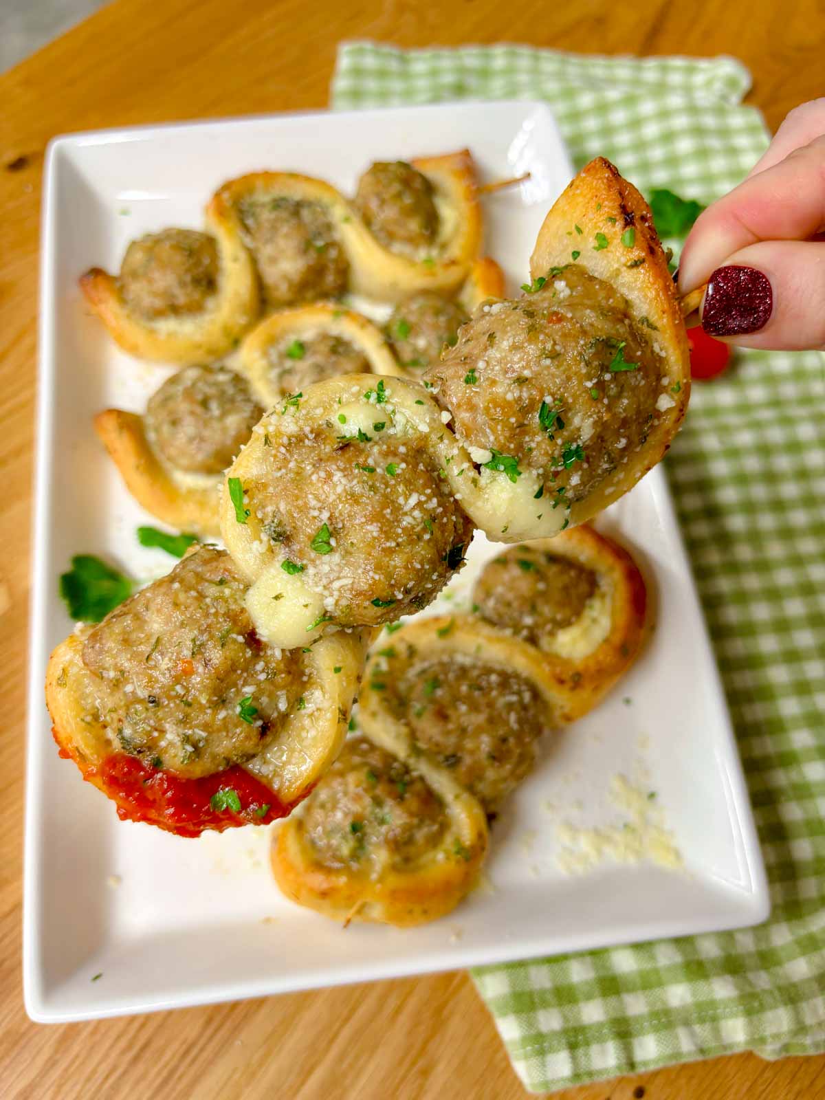 a white plate has chicken parmesan meatball subs on a stick with a side of marinara sauce to dip. garnished with parsley and parmesan cheese. one is held close to camera.