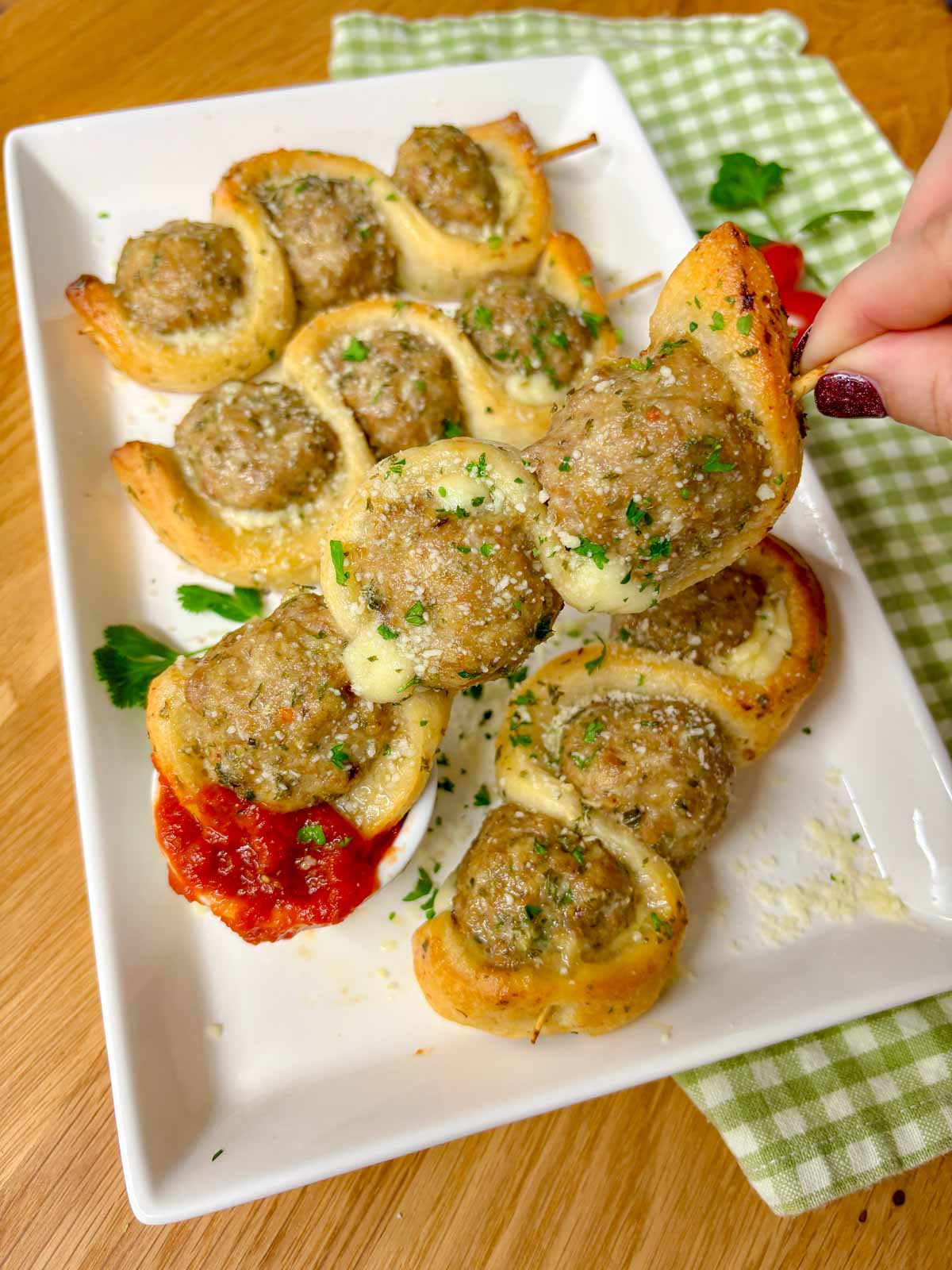 a white plate has chicken parmesan meatball subs on a stick with a side of marinara sauce to dip. garnished with parsley and parmesan cheese. one is dipped in the sauce