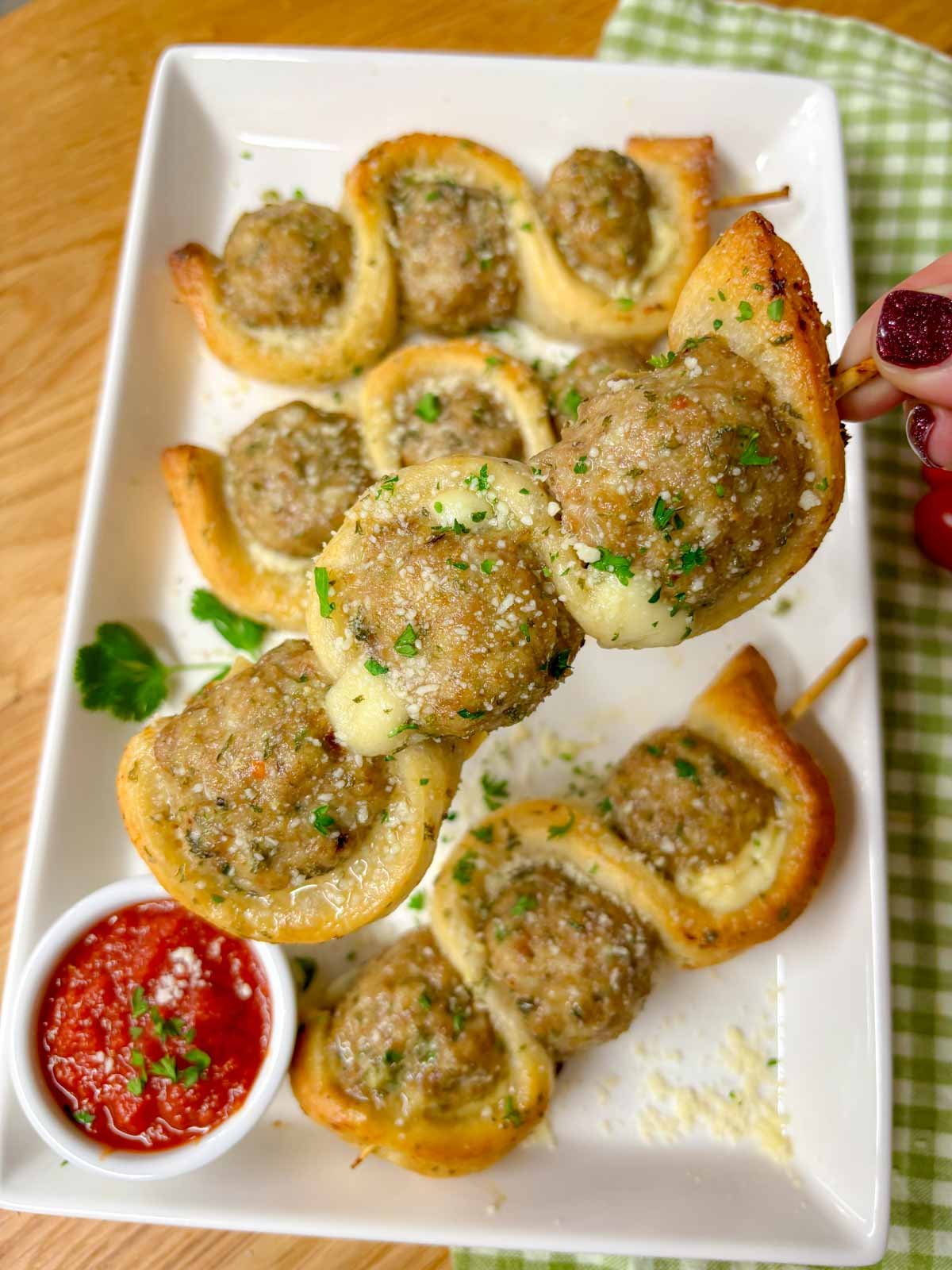 a white plate has chicken parmesan meatball subs on a stick with a side of marinara sauce to dip. garnished with parsley and parmesan cheese. one is held up close to camera.
