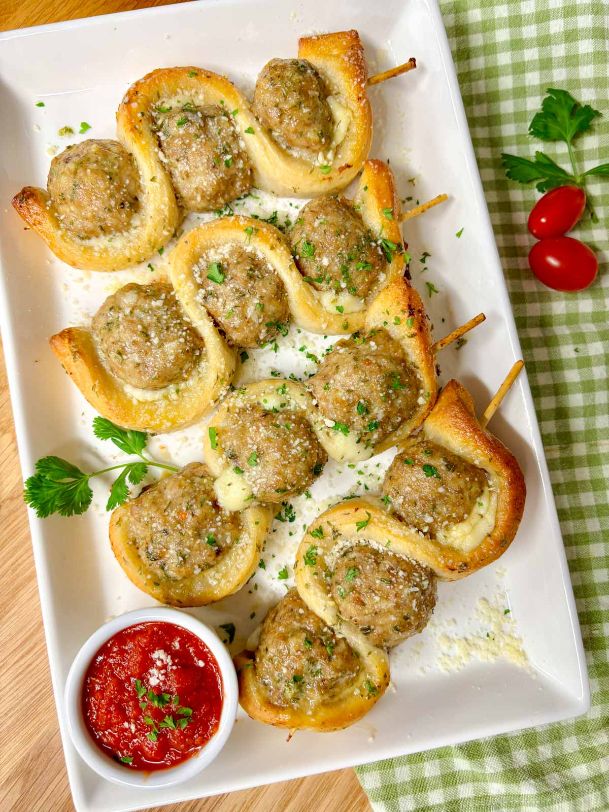 a white plate has chicken parmesan meatball subs on a stick with a side of marinara sauce to dip. garnished with parsley and parmesan cheese.