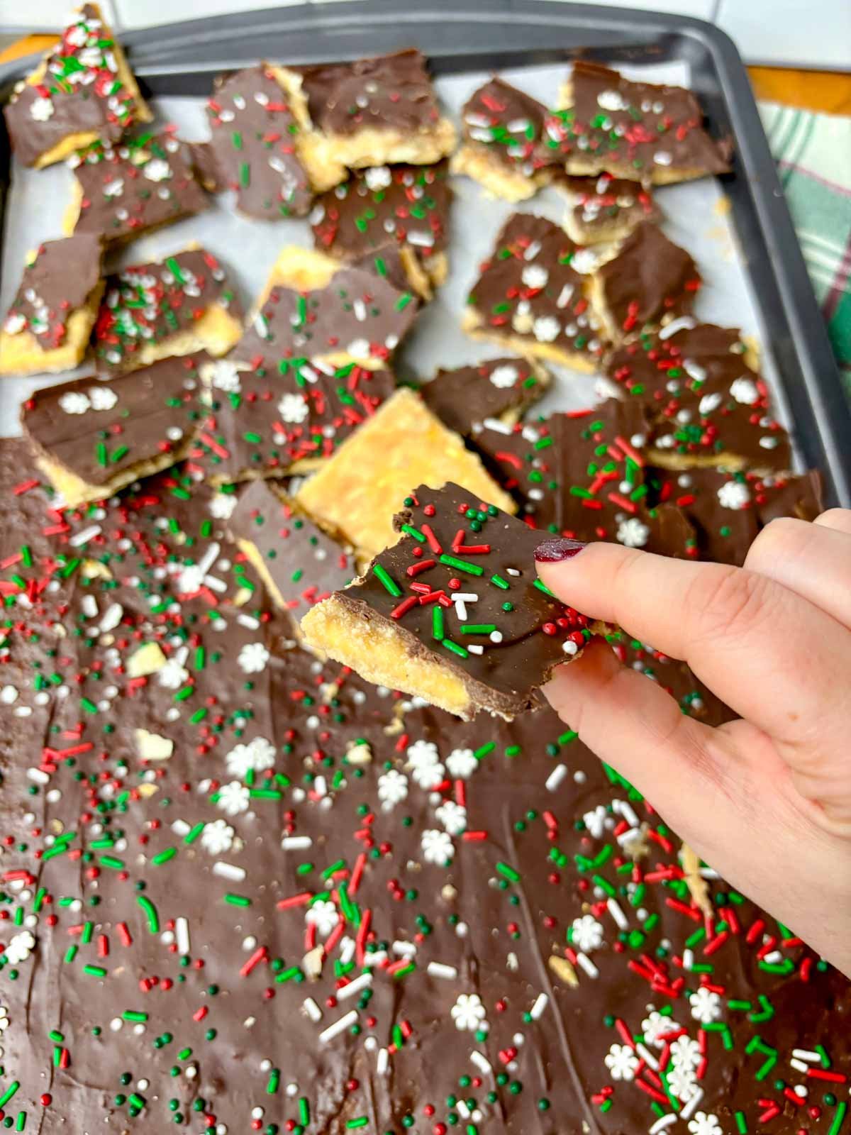 a sheet pan is filled with pieces of cracker toffee (Christmas crack). one piece is held close to camera