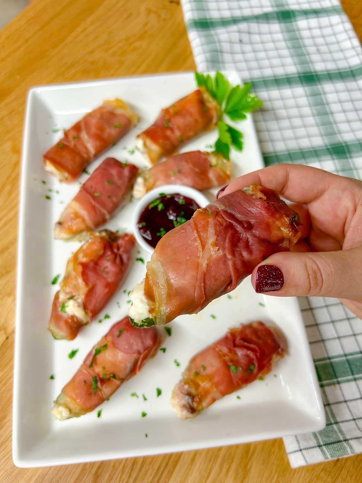 a white platter with  prosciutto wrapped cranberry brie jalapeno poppers garnished with chopped parsley. one is held close to camera