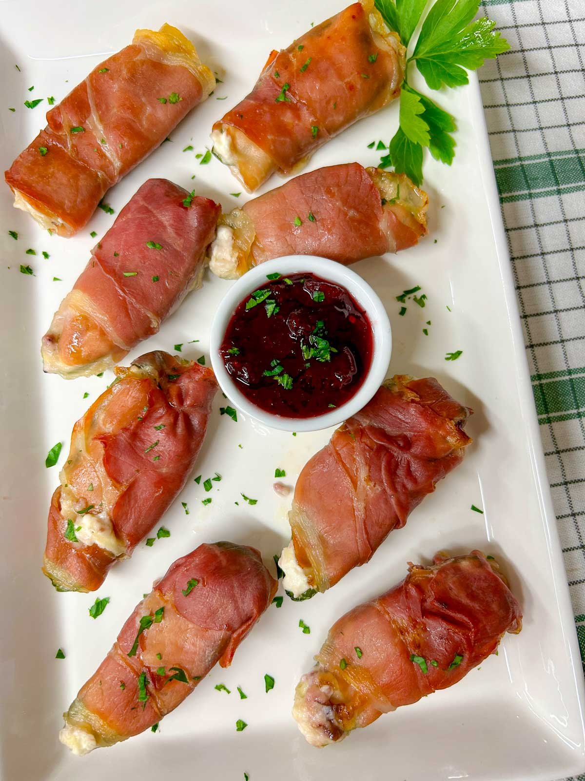 a white platter with  prosciutto wrapped cranberry brie jalapeno poppers garnished with chopped parsley.