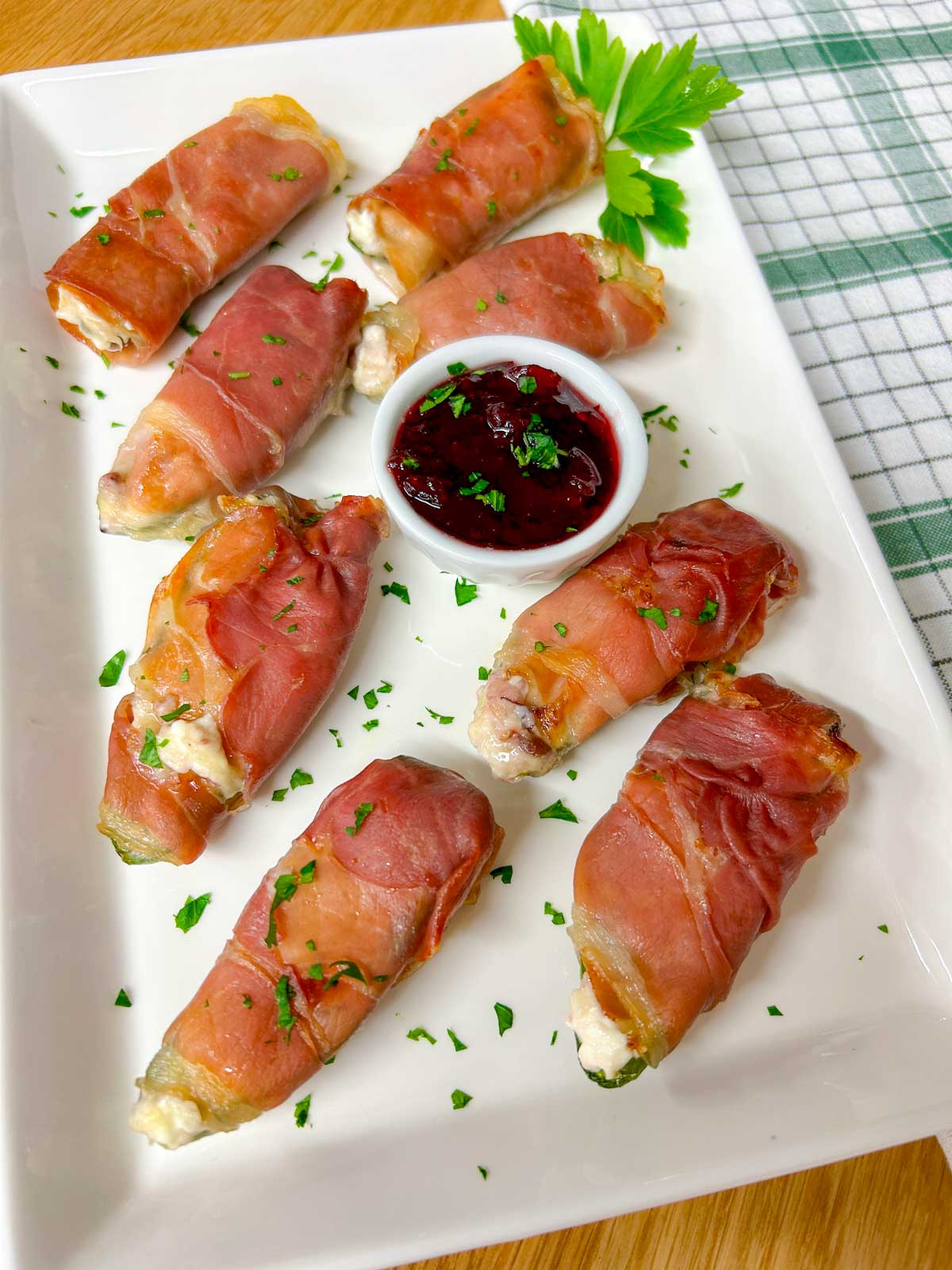 a white platter with  prosciutto wrapped cranberry brie jalapeno poppers garnished with chopped parsley.