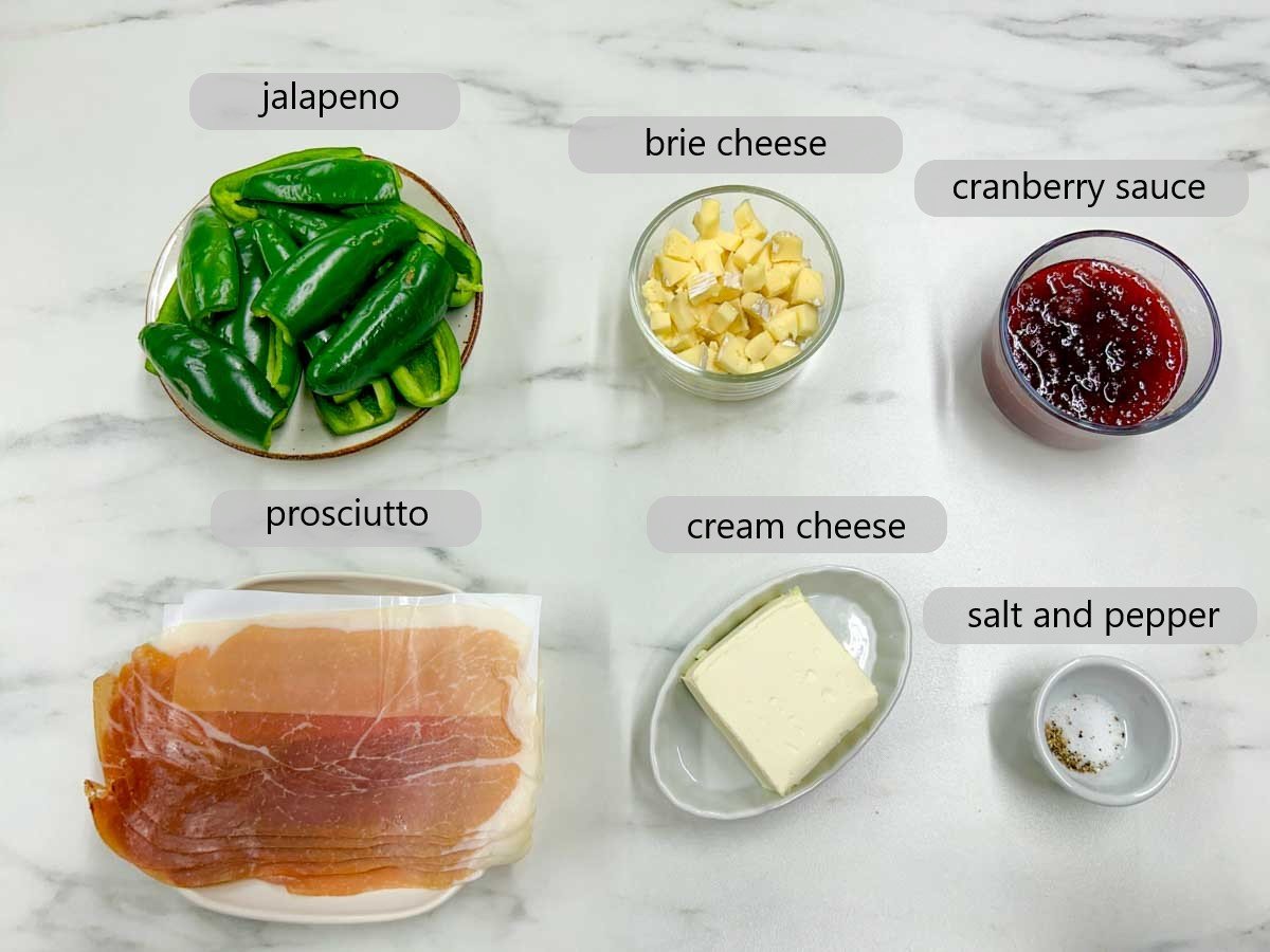 a kitchen counter with bowls of ingredients to make cranberry brie jalapeno poppers with text overlays reading jalapeno, brie cheese, cranberry sauce, prosciutto, cream cheese, salt and pepper.