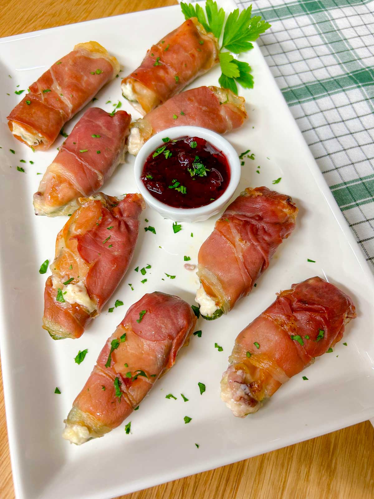 a white platter with  prosciutto wrapped cranberry brie jalapeno poppers garnished with chopped parsley.