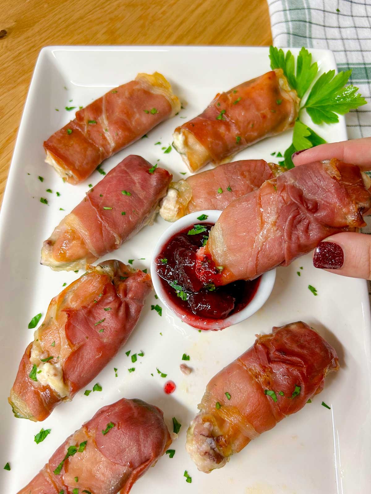 a white platter with  prosciutto wrapped cranberry brie jalapeno poppers garnished with chopped parsley. one is dipped in cranberry sauce