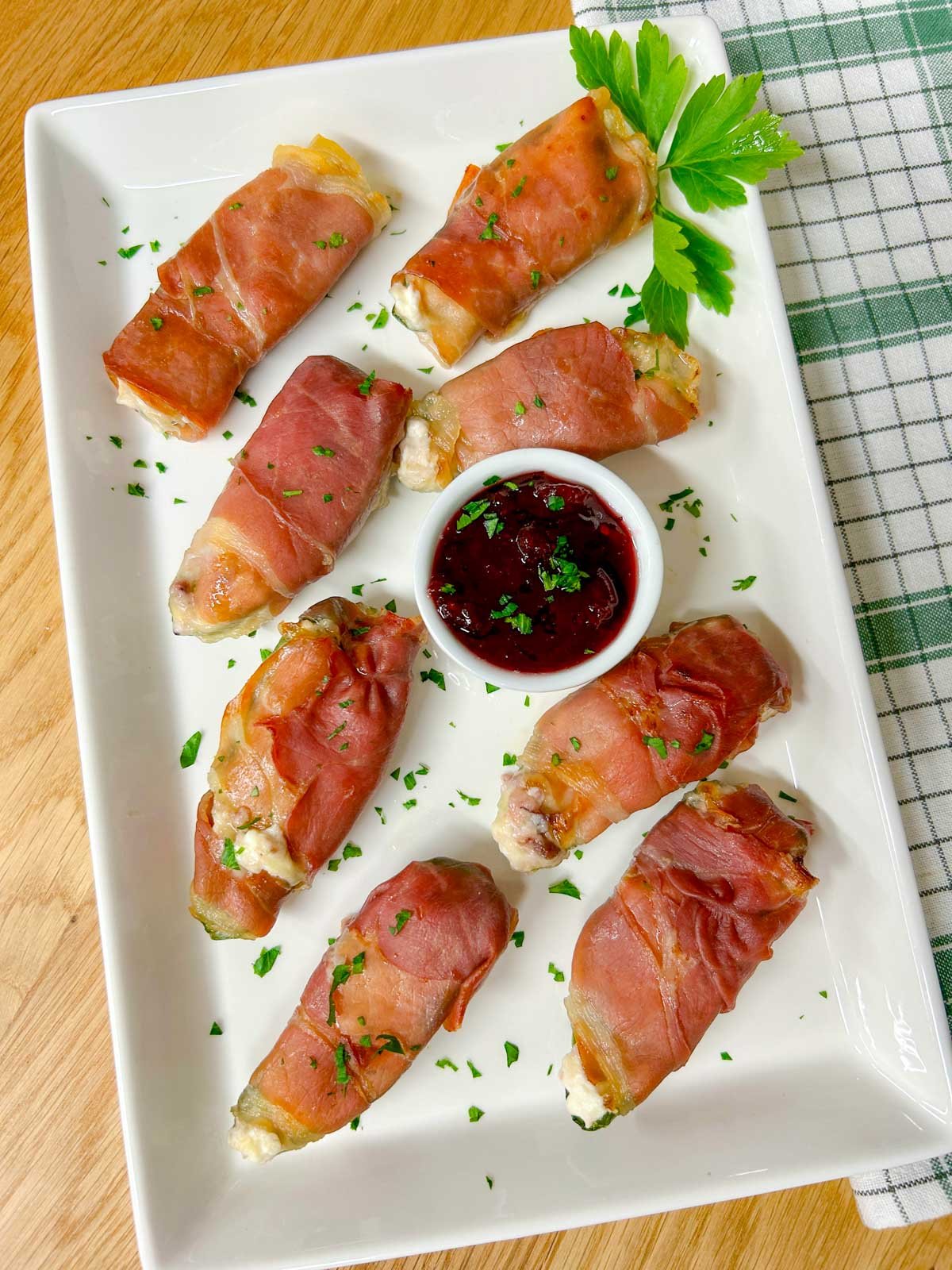 a white platter with  prosciutto wrapped cranberry brie jalapeno poppers garnished with chopped parsley.