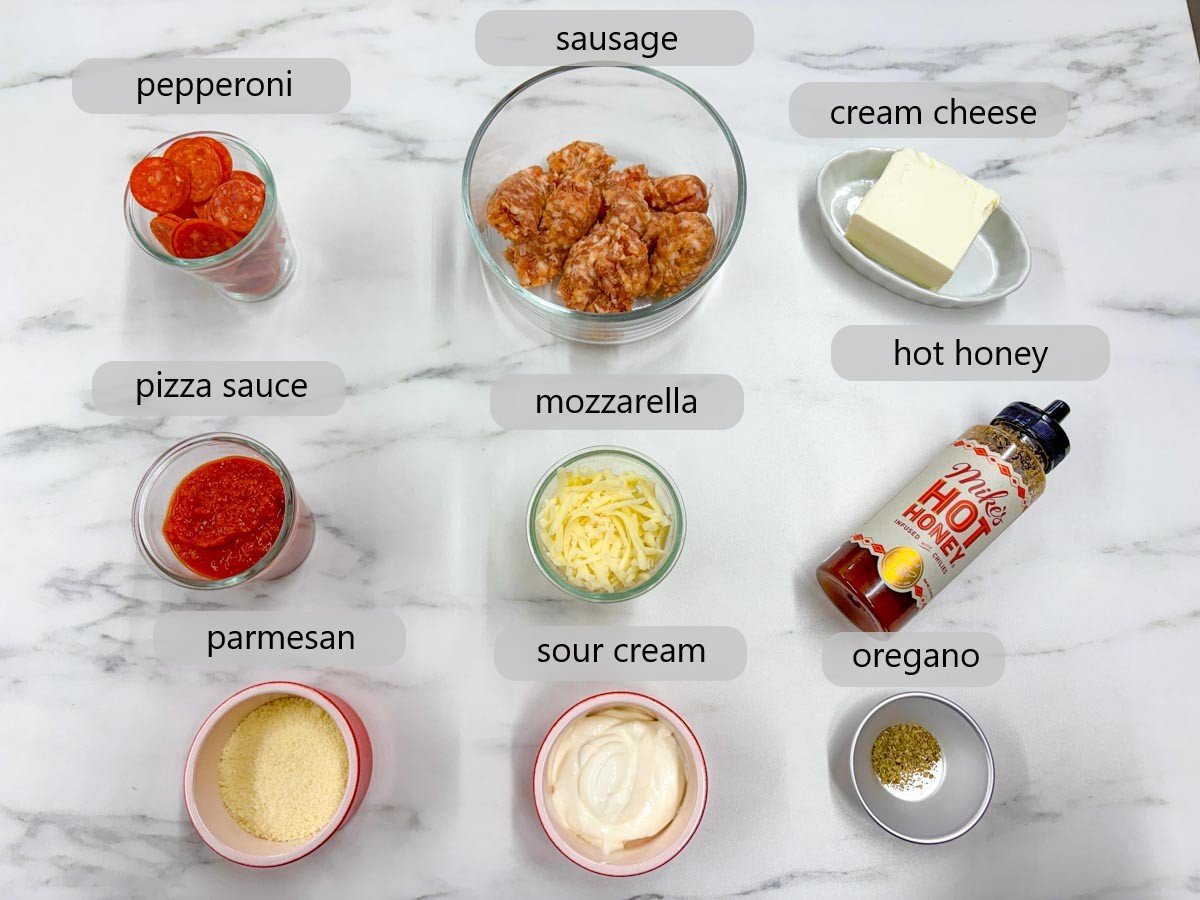 kitchen counter with ingredients and text overlays reading pepperoni, sausage, cream cheese, pizza sauce, mozzarella, hot honey, parmesan, sour cream and oregano.