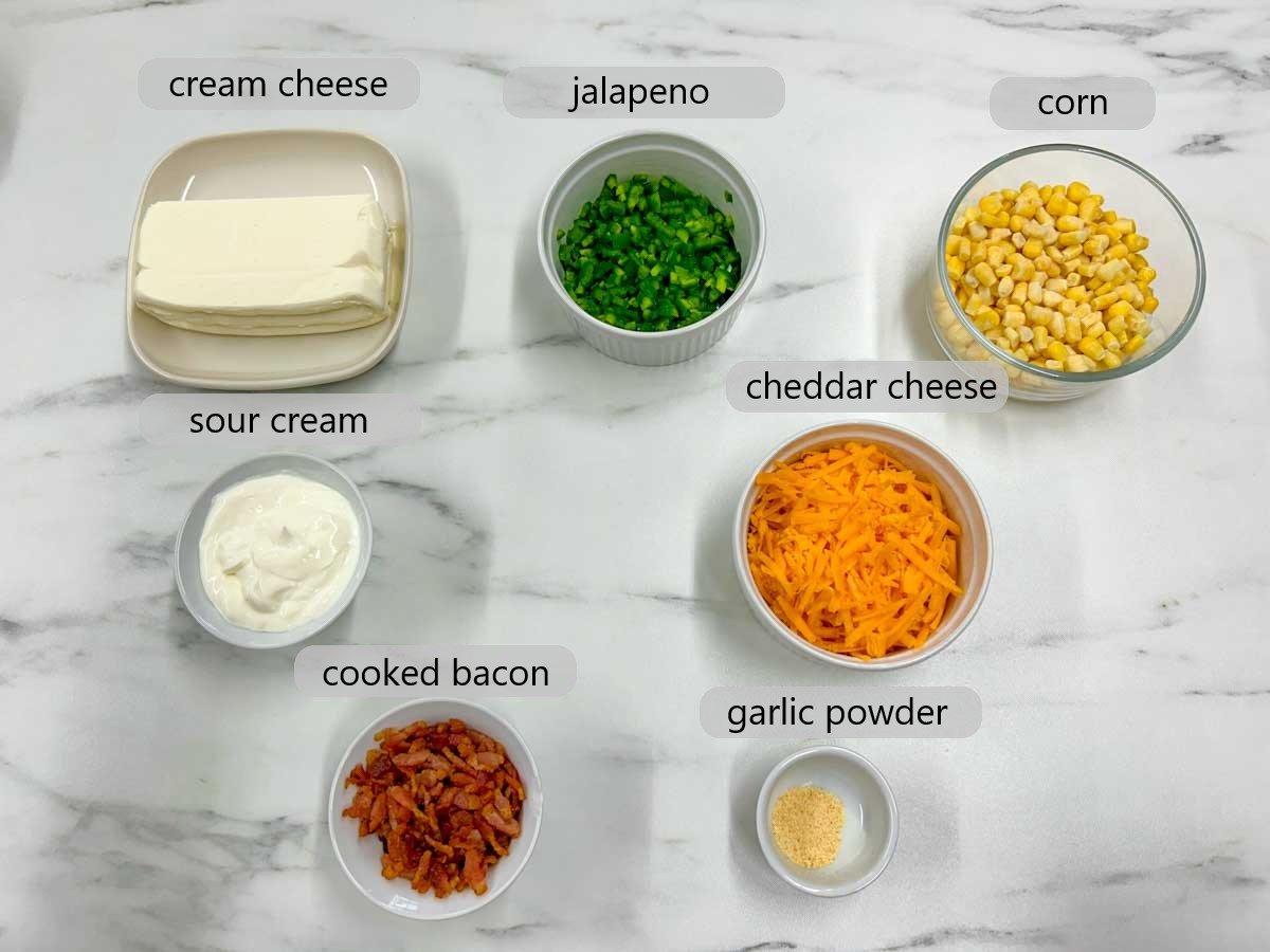 kitchen counter with bowls of ingredients and text overlays reading cream cheese, jalapeno, corn, sour cream, cheddar cheese, cooked bacon, and garlic powder.