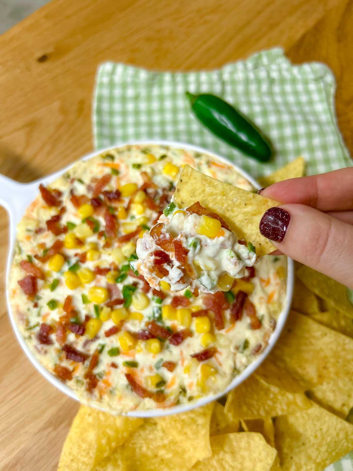 a baking dish is filled with jalapeno popper corn dip and garnished with jalapenos, bacon and corn.  one chip is dipped and held close to camera