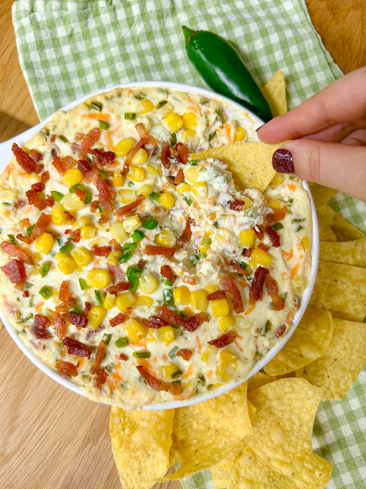 a baking dish is filled with jalapeno popper corn dip and garnished with jalapenos, bacon and corn. a tortilla chip is dipped in