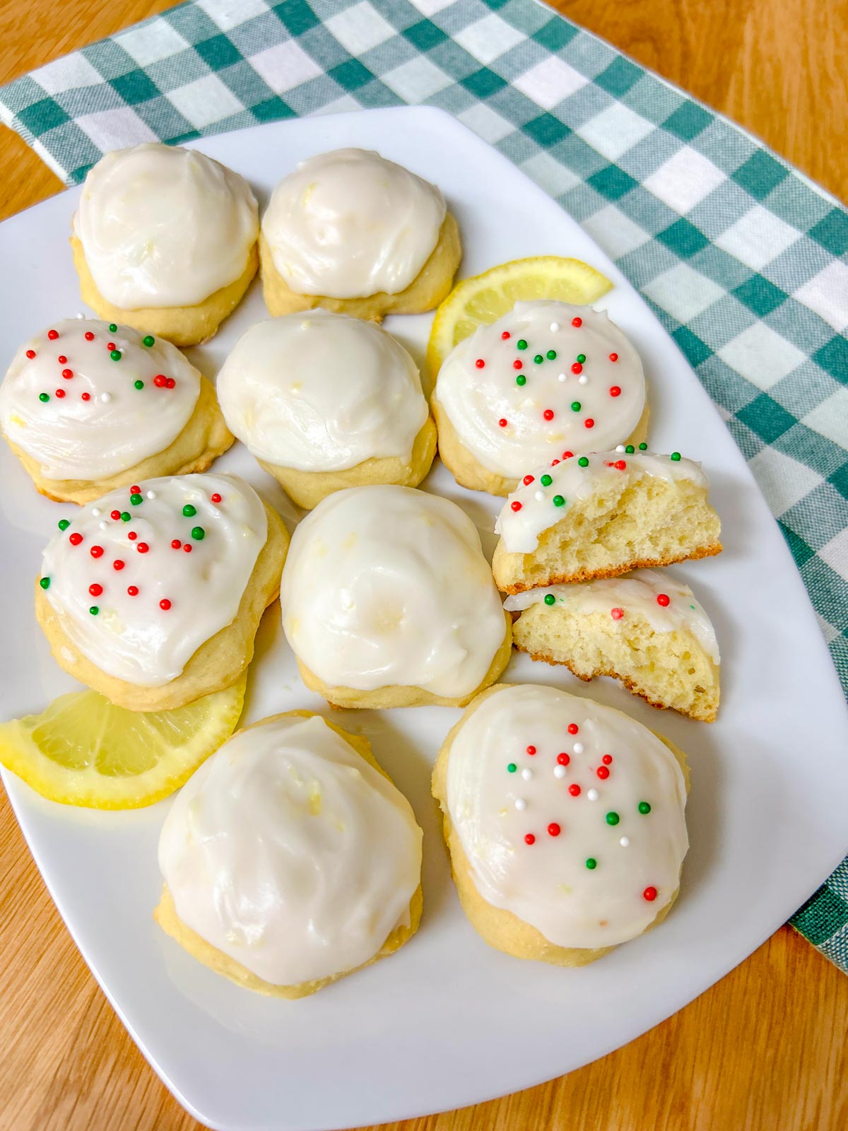a white plate is filled with Italian lemon ricotta cookies, some have sprinkles and lemons garnish the plate. one is cut in half
