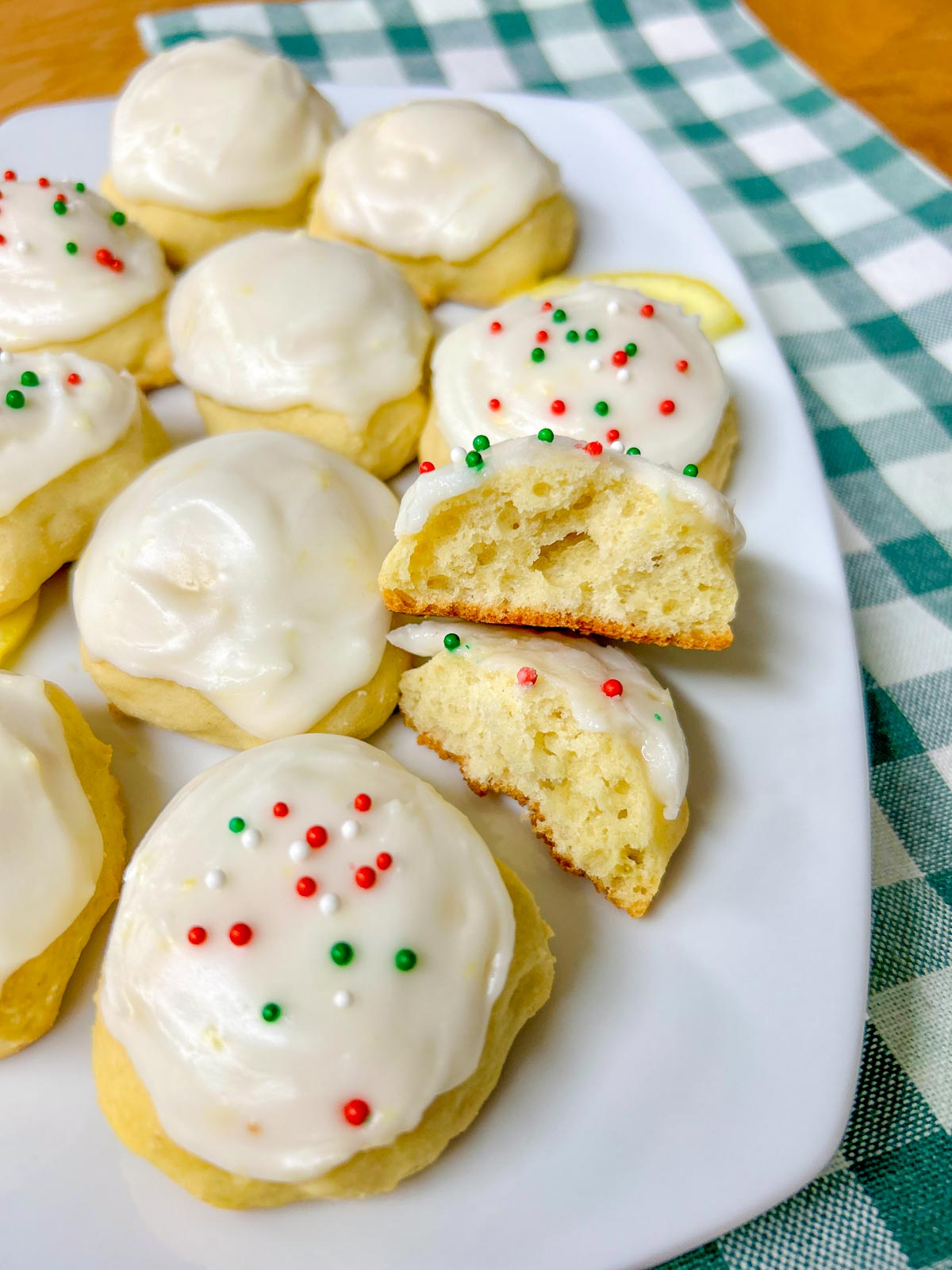 a white plate is filled with Italian lemon ricotta cookies, some have sprinkles and lemons garnish the plate. one is cut in half