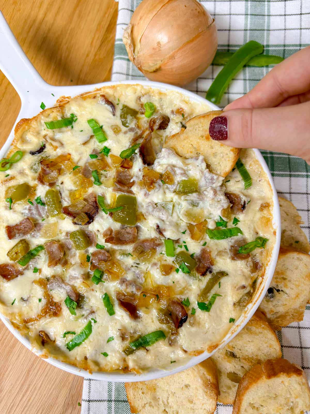 a white baking dish is filled with Philly cheesesteak dip. peppers, onions, roast beef and green onions garnish the dip. a piece of baguette is dipped