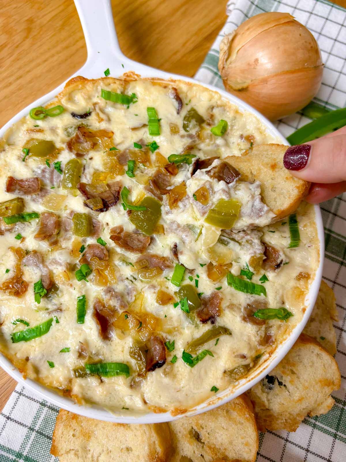 a white baking dish is filled with Philly cheesesteak dip. peppers, onions, roast beef and green onions garnish the dip. a piece of baguette is dipped