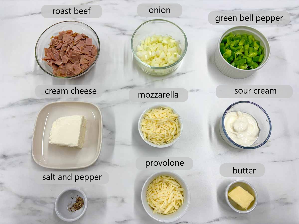kitchen counter with ingredients in bowls and text overlay reading roast beef, onion, bell pepper, cream cheese, mozzarella, sour cream, salt and pepper, provolone and butter.