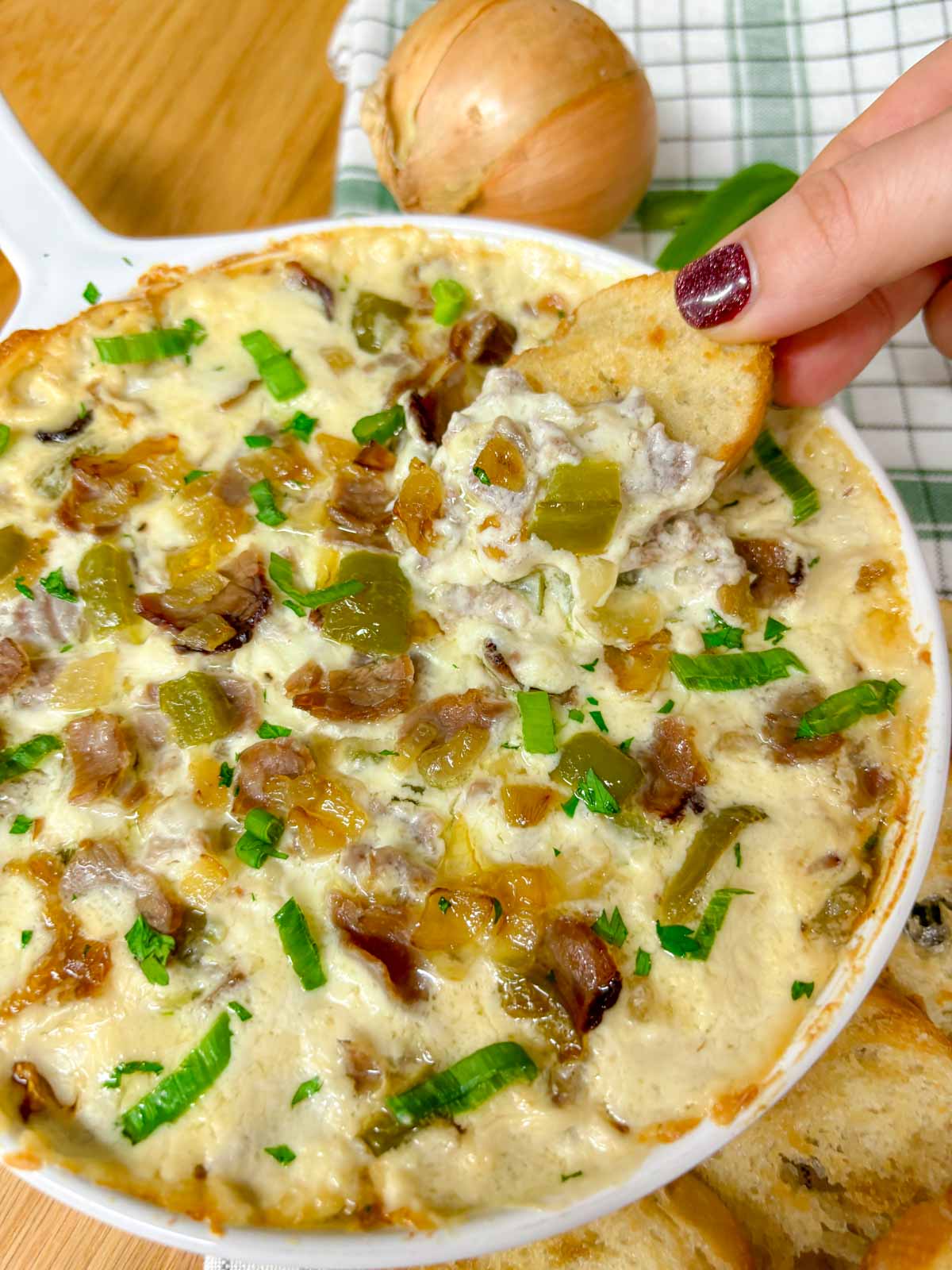 a white baking dish is filled with Philly cheesesteak dip. peppers, onions, roast beef and green onions garnish the dip. a piece of baguette is dipped in
