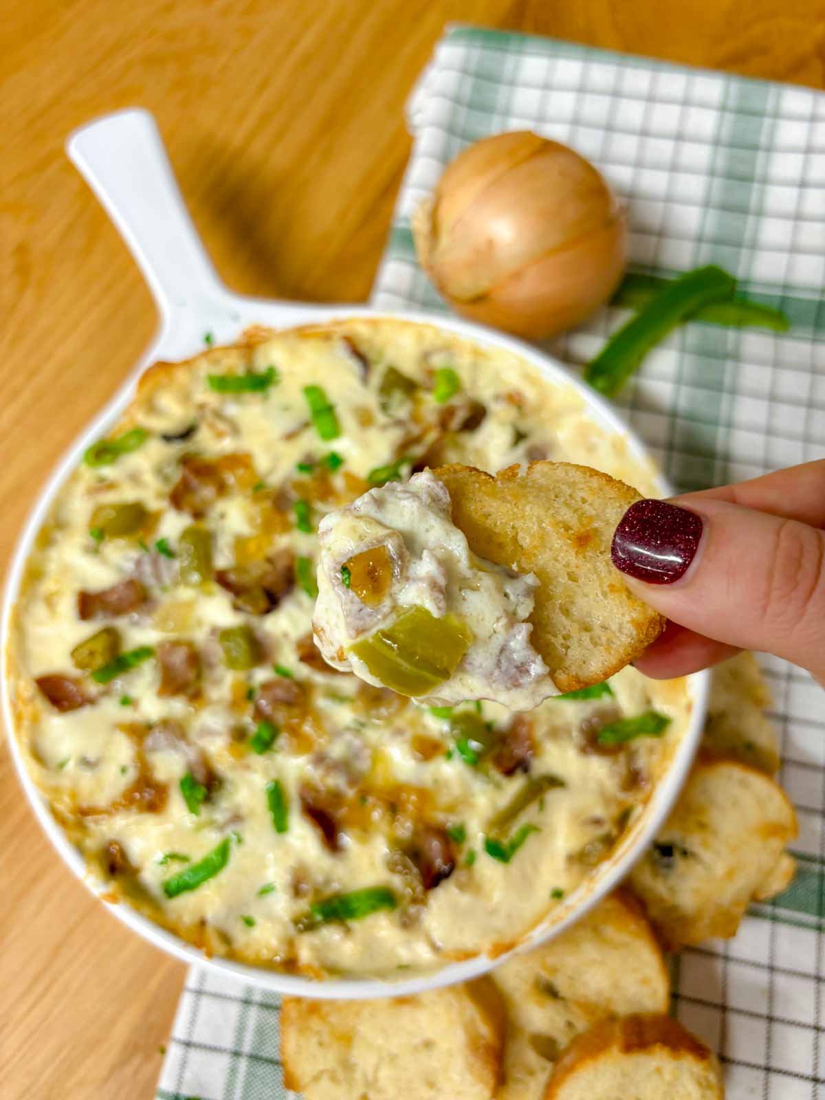 a white baking dish is filled with Philly cheesesteak dip. peppers, onions, roast beef and green onions garnish the dip. a piece of baguette is dipped