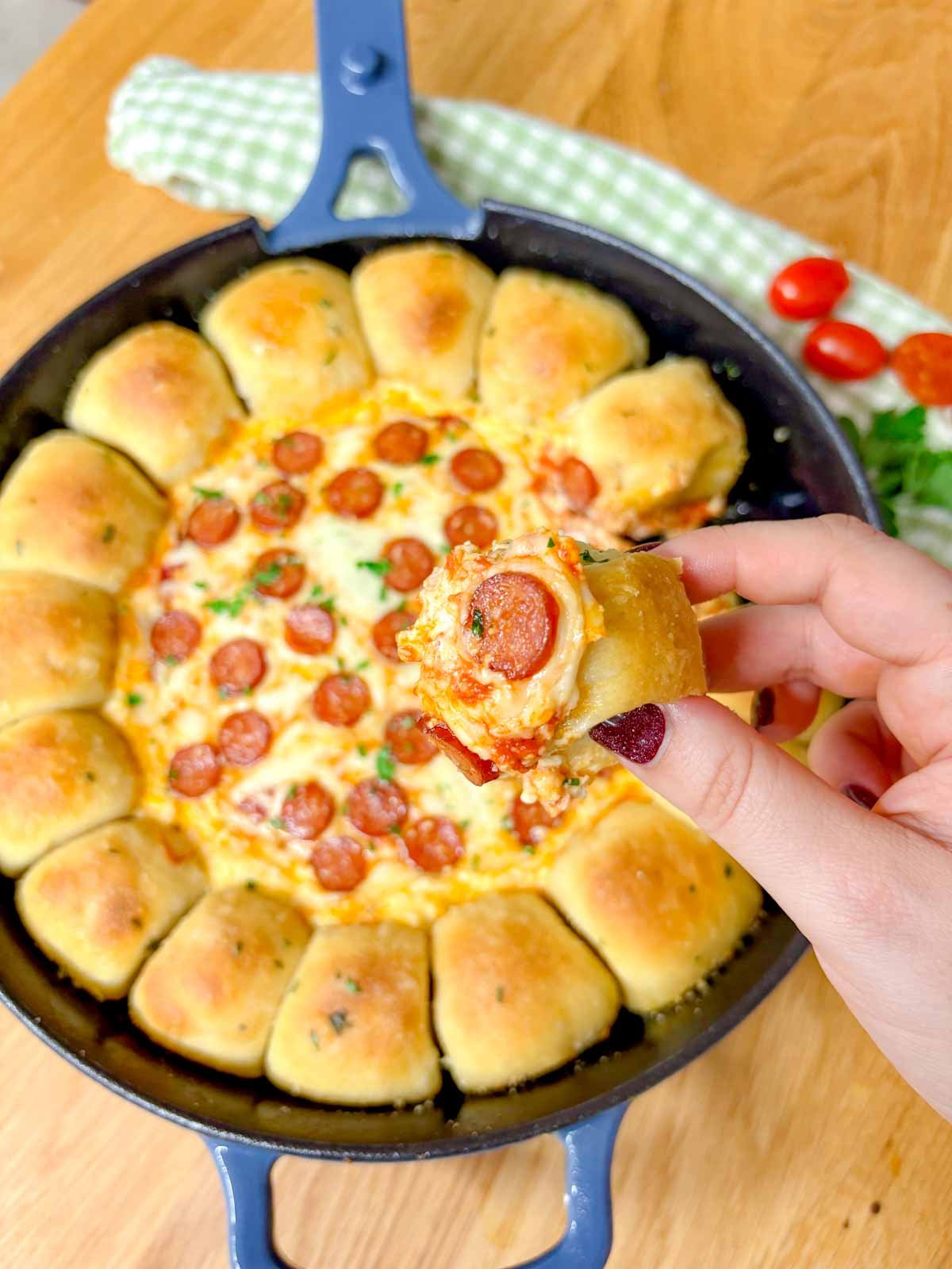 a pizza dip ring with pizza dough balls and a pizza dip in the middle is in a cast iron skillet. one piece of dipped bread is held close to camera