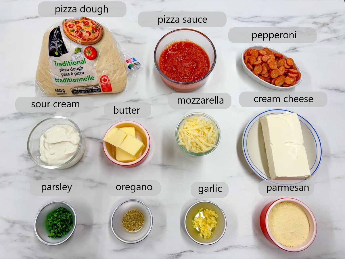 counter with ingredients and text overlays reading pizza dough, pizza sauce, pepperoni, sour cream, butter, mozzarella, cream cheese, parsley, oregano, garlic and parmesan.