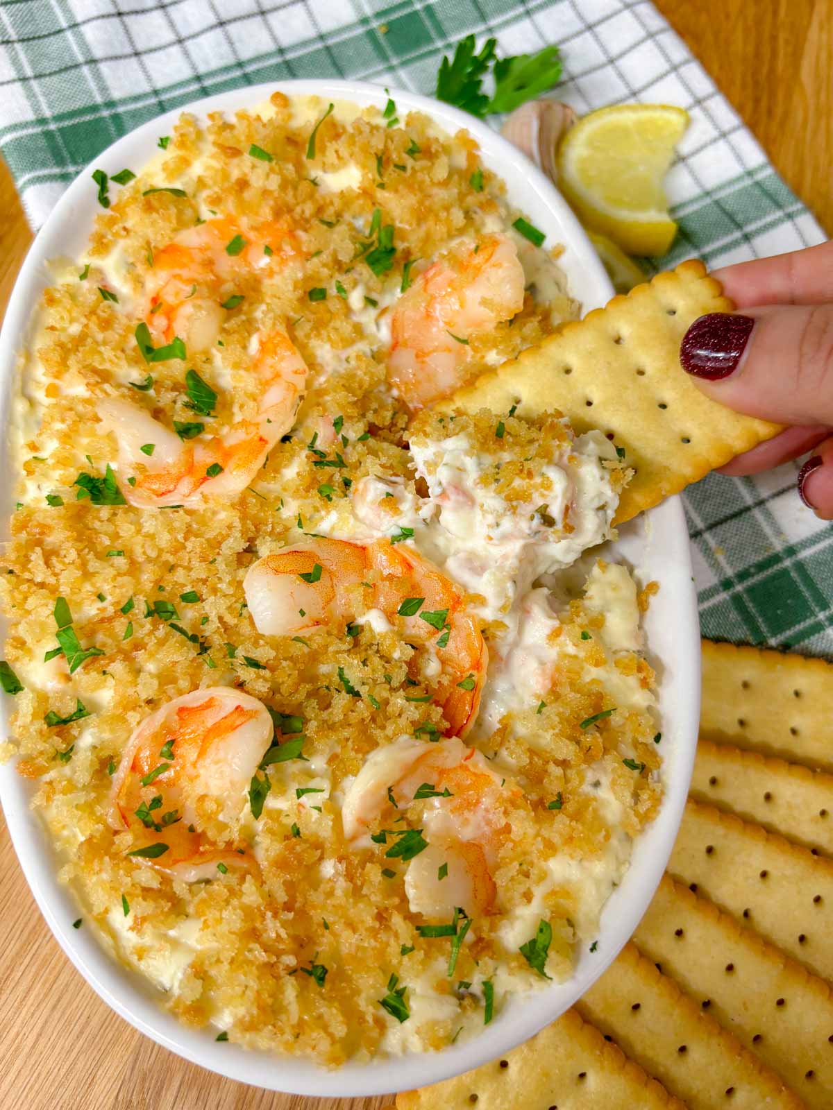 a white baking dish is filled with shrimp dip and garnished with breadcrumbs and parsley. a cracker is dipped in