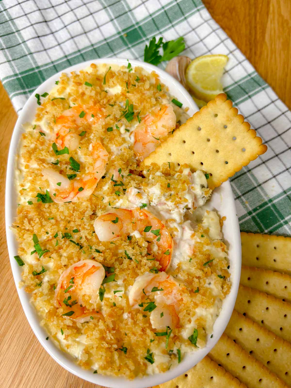 a white baking dish is filled with shrimp scampi dip and garnished with breadcrumbs and parsley. a cracker is dipped