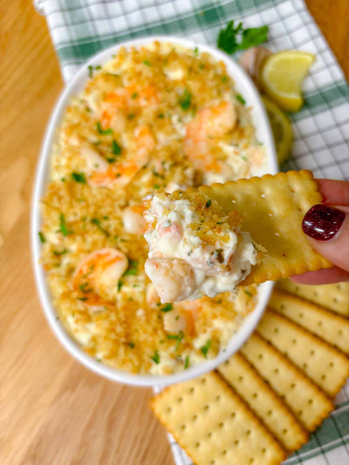 a white baking dish is filled with shrimp scampi dip and garnished with breadcrumbs and parsley. a cracker is dipped and held close to camera