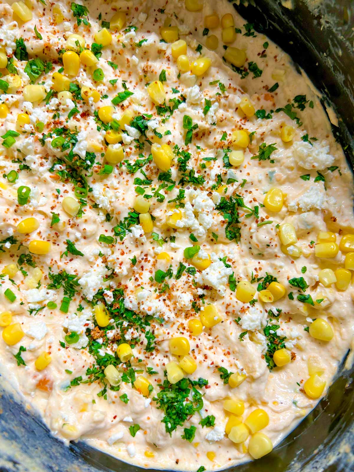 a slow cooker is filled with street corn dip and garnished with cilantro, corn and cotija cheese.