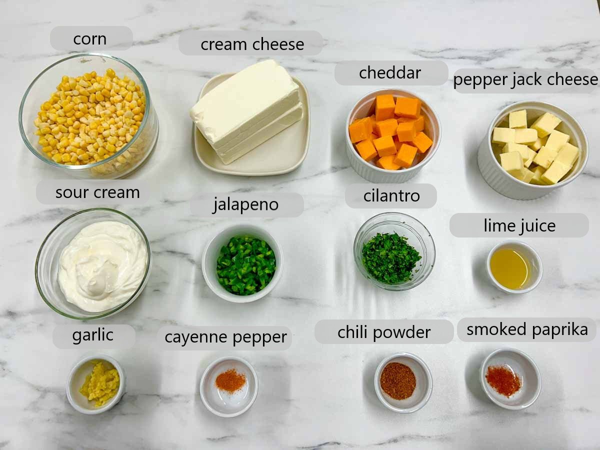 a countertop with bowls of ingredients to make street corn dip and text overlays reading corn, cream cheese, cheddar, pepper jack cheese, sour cream, jalapeno, cilantro, lime juice, garlic, cayenne pepper, chili powder and smoked paprika