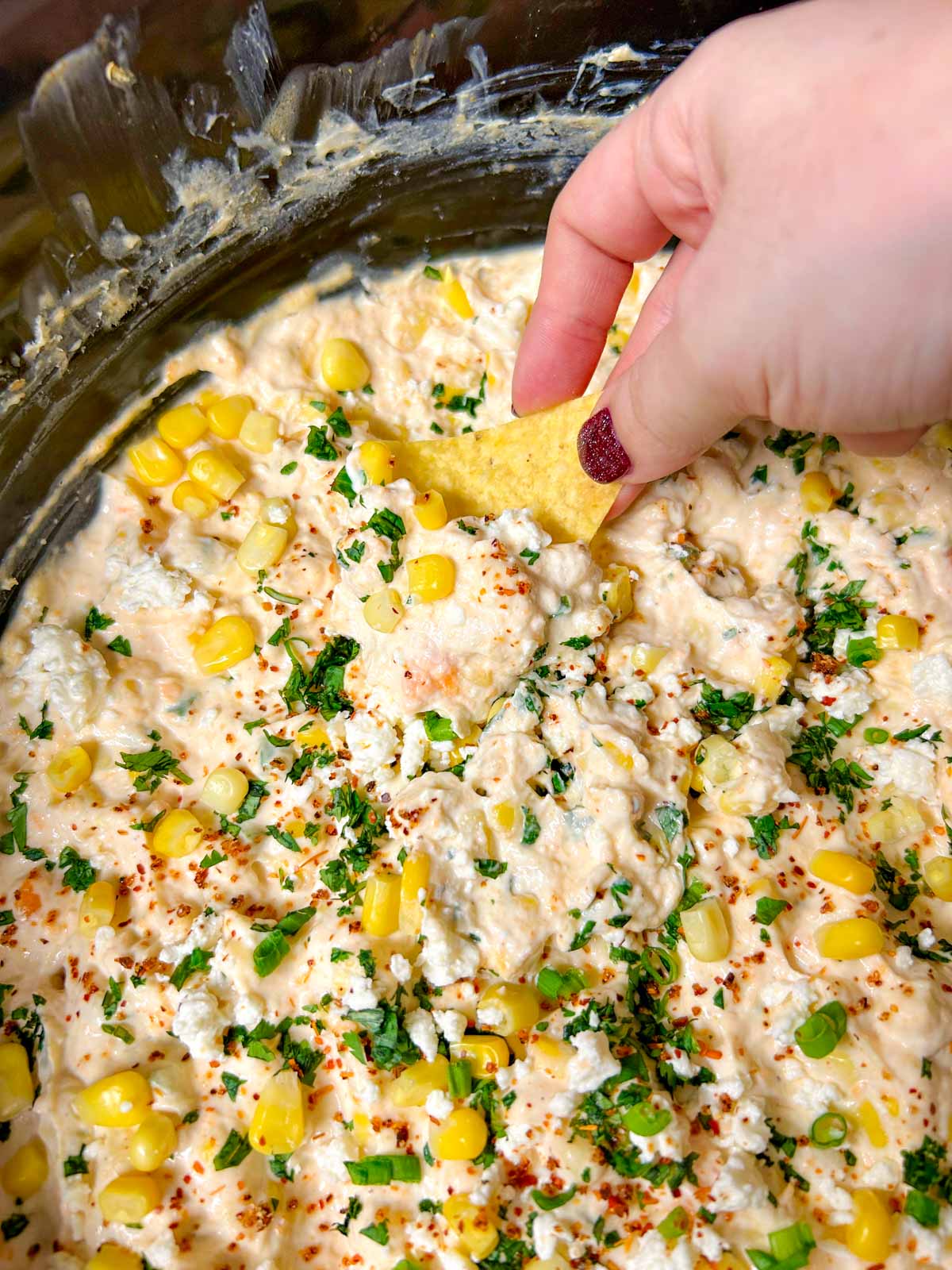 a slow cooker is filled with street corn dip and garnished with cilantro, corn and cotija cheese. a chip is dipped