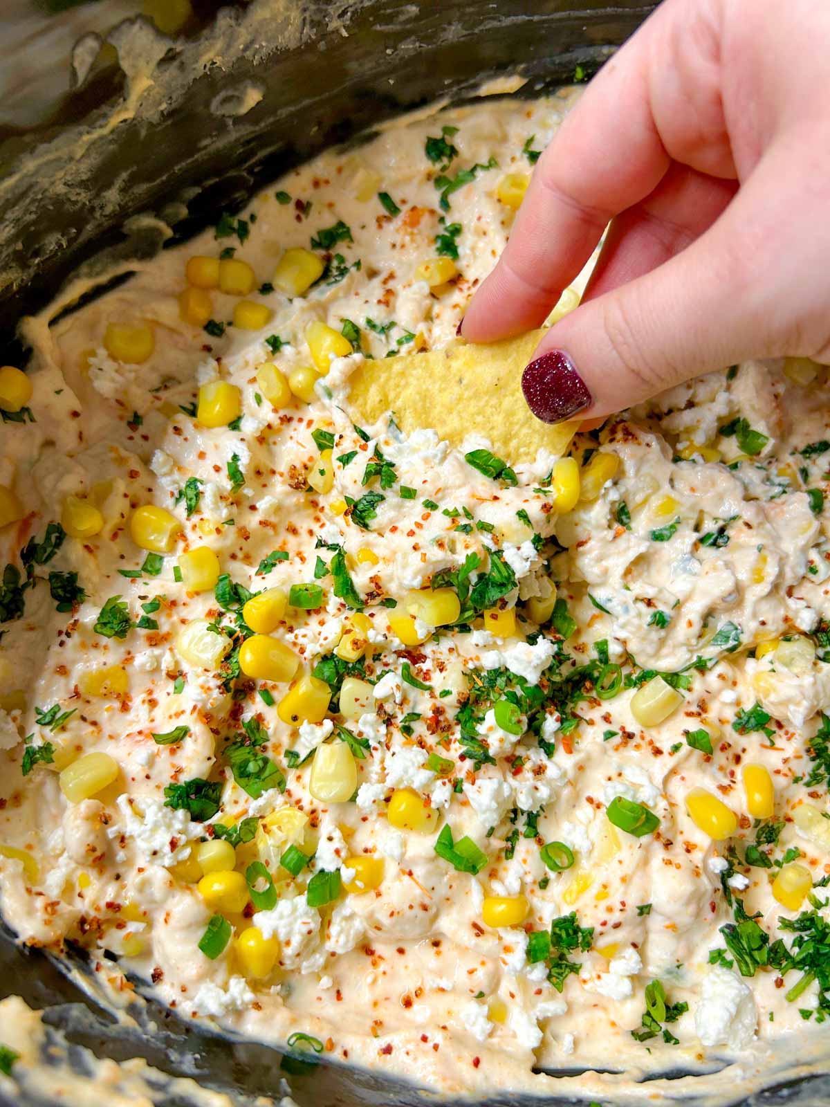 a slow cooker is filled with street corn dip and garnished with cilantro, corn and cotija cheese. a chip is dipped