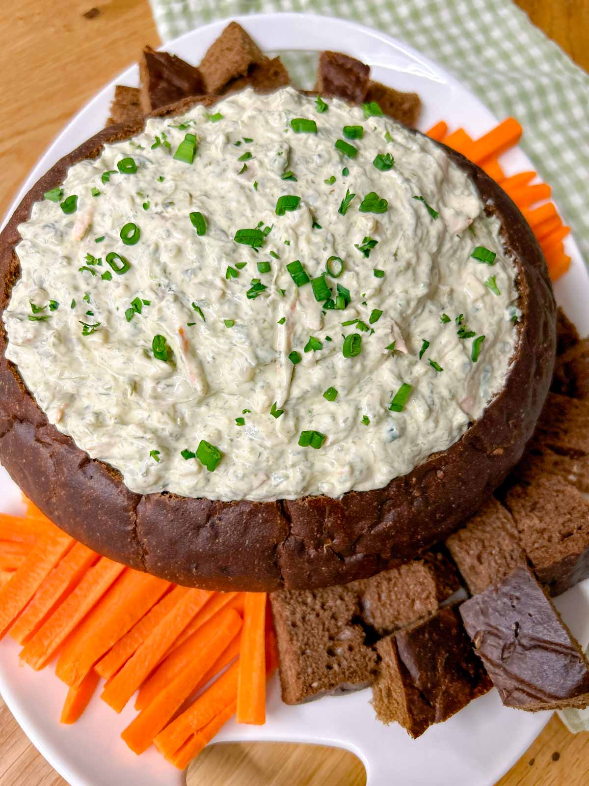 a pumpernickel bread bowl is filled with spinach dip and served with bread and carrot sticks to dip.