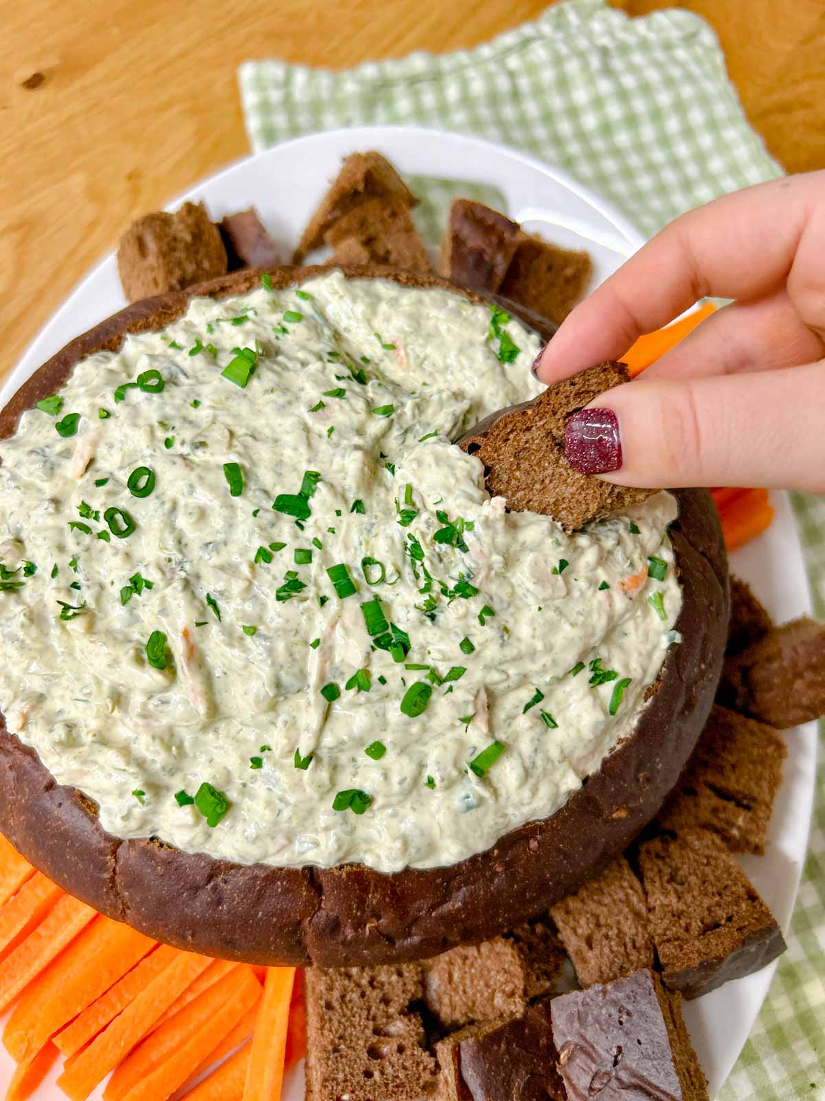 a pumpernickel bread bowl is filled with spinach dip and served with bread and carrot sticks to dip. a piece of bread is dipped in