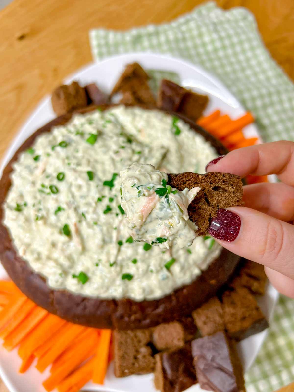 a pumpernickel bread bowl is filled with spinach dip and served with bread and carrot sticks to dip. a piece of bread is dipped and held close to camera