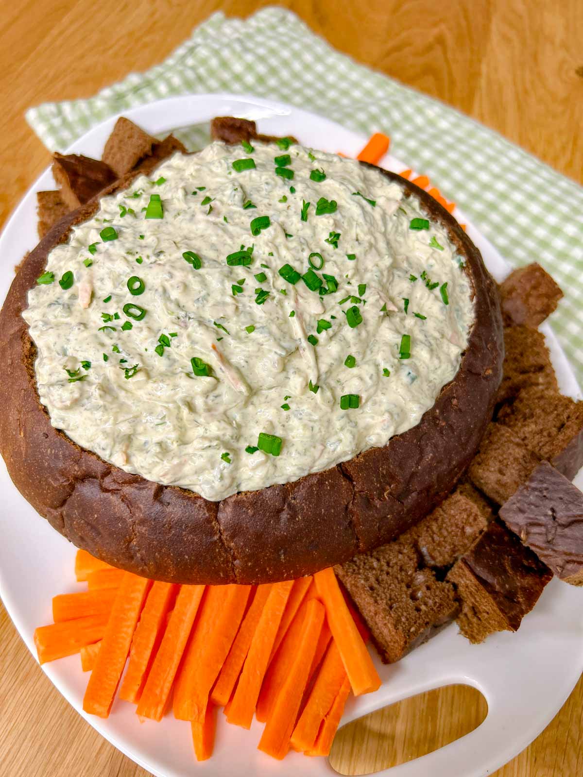 a pumpernickel bread bowl is filled with spinach dip and served with bread and carrot sticks to dip.
