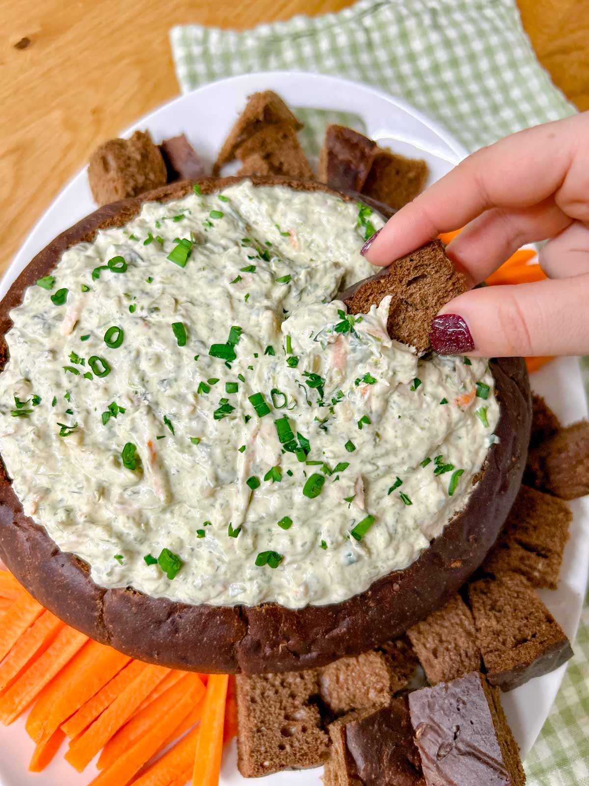 a pumpernickel bread bowl is filled with spinach dip and served with bread and carrot sticks to dip. a piece of bread is dipped