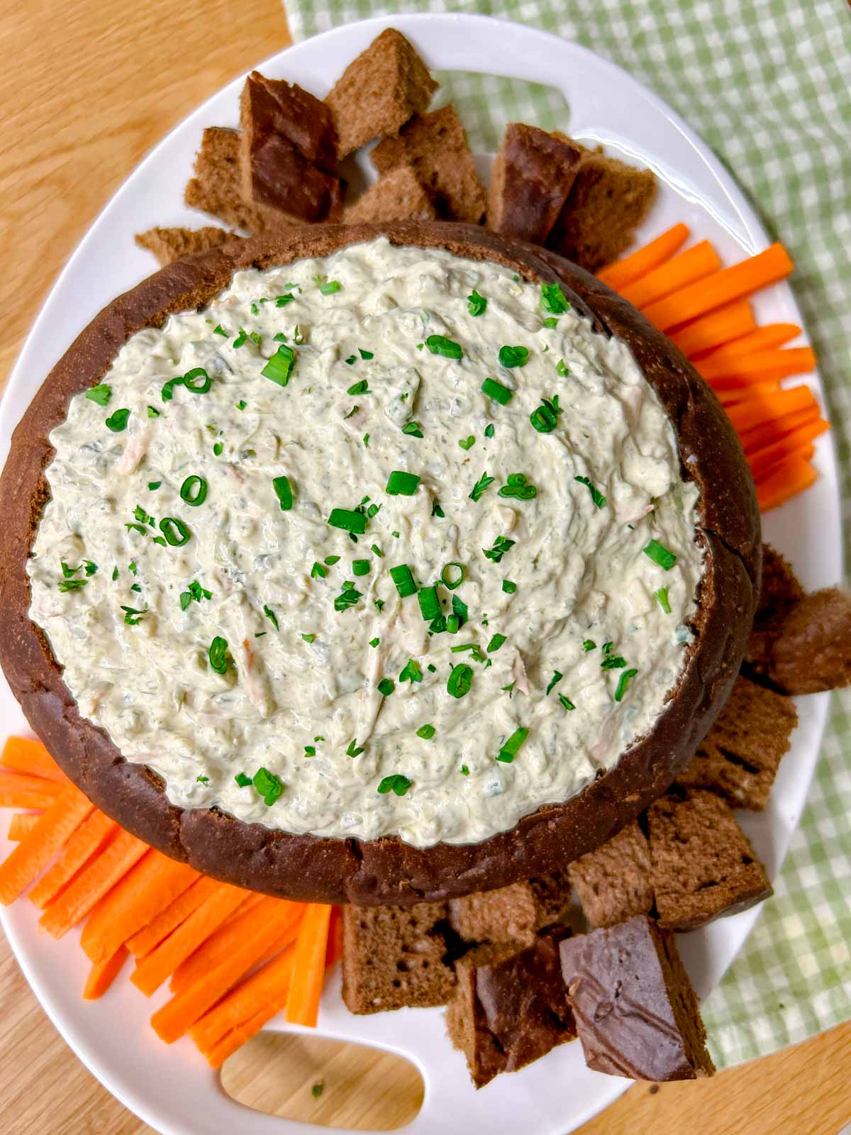 a pumpernickel bread bowl is filled with spinach dip and served with bread and carrot sticks to dip.