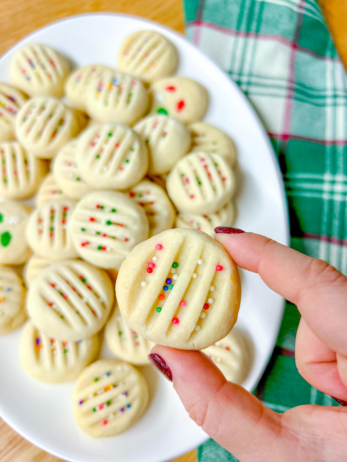 a white platter is filled with whipped shortbread cookies that are topped with sprinkles. one is held close to camera