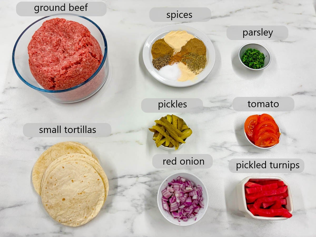 kitchen counter with bowls of ingredients and text overlay reading ground beef, spices, parsley, small tortillas, pickles, tomato, red onion, and pickled turnips.