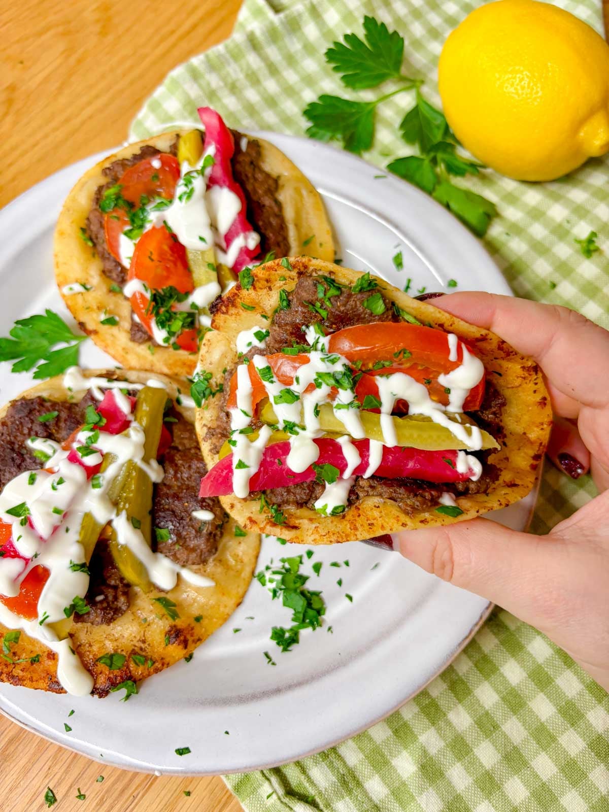 a beef kebab smash taco is filled with tomato, pickle, pickled turnip, garlic sauce, and parsley and is held up close to camera