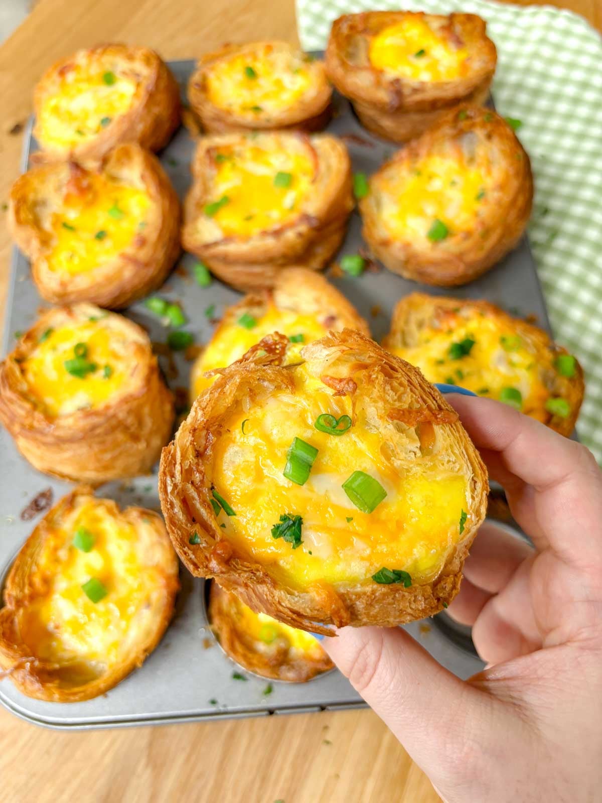 a muffin tin is filled with breakfast croissant cups and garnished with green onions and parsley. one is held up.