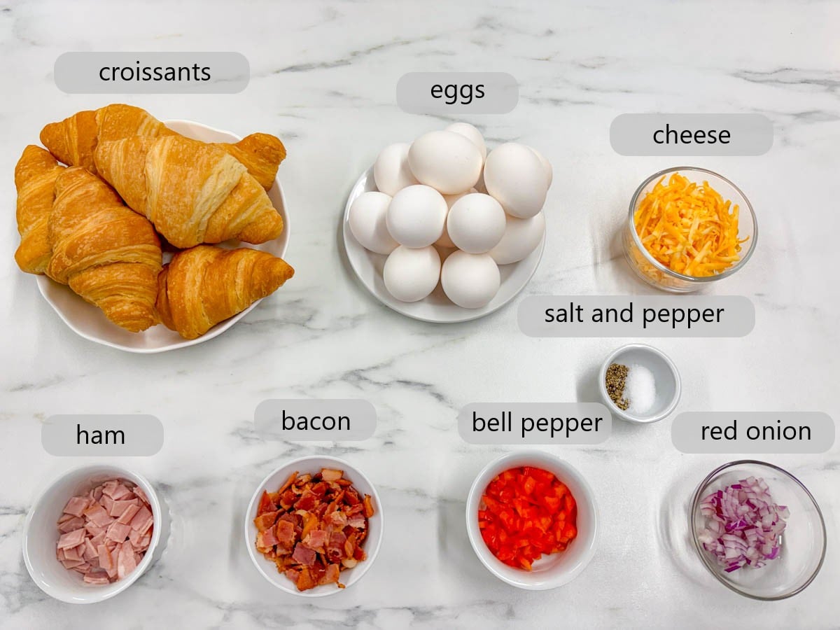 kitchen counter with bowls of ingredients and text overlays reading croissants, eggs, cheese, salt and pepper, ham, bacon, bell pepper, and red onion.