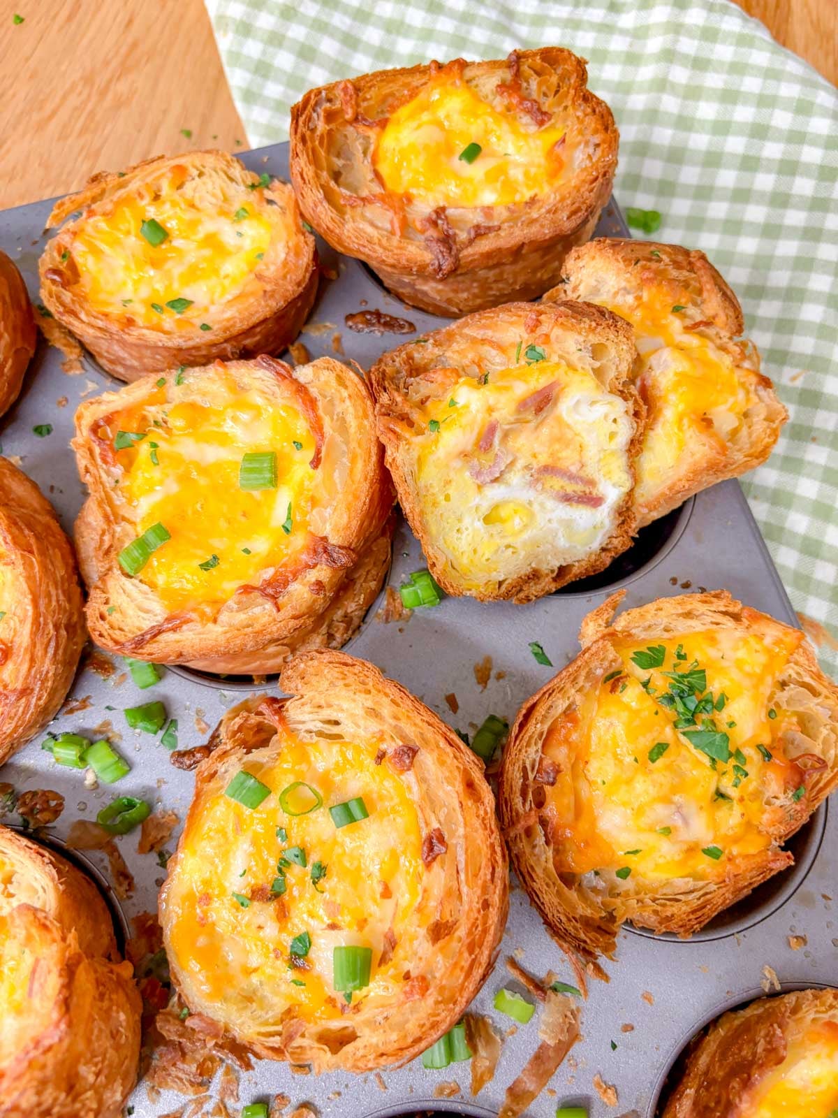 a muffin tin is filled with breakfast croissant cups and garnished with green onions and parsley. one is cut in half