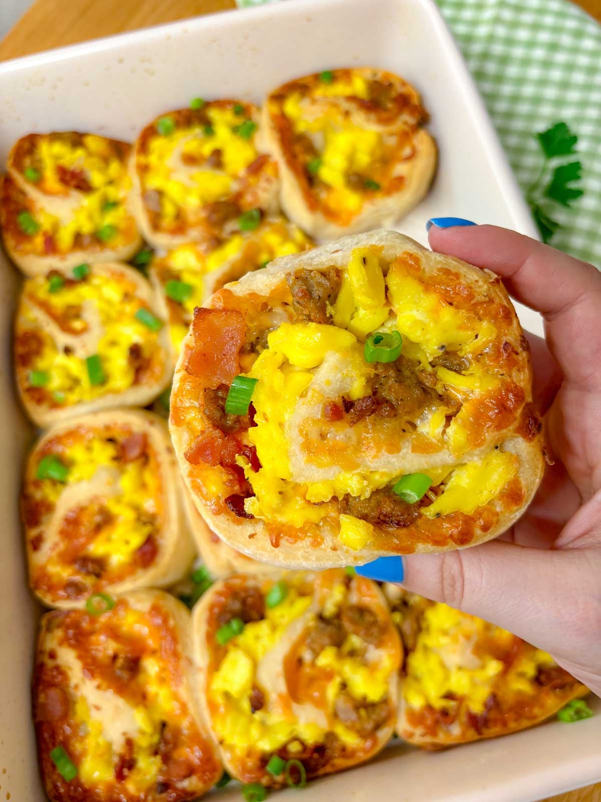 a sheet pan is filled with baked breakfast rolls that are garnished with green onion. one is held close to the camera