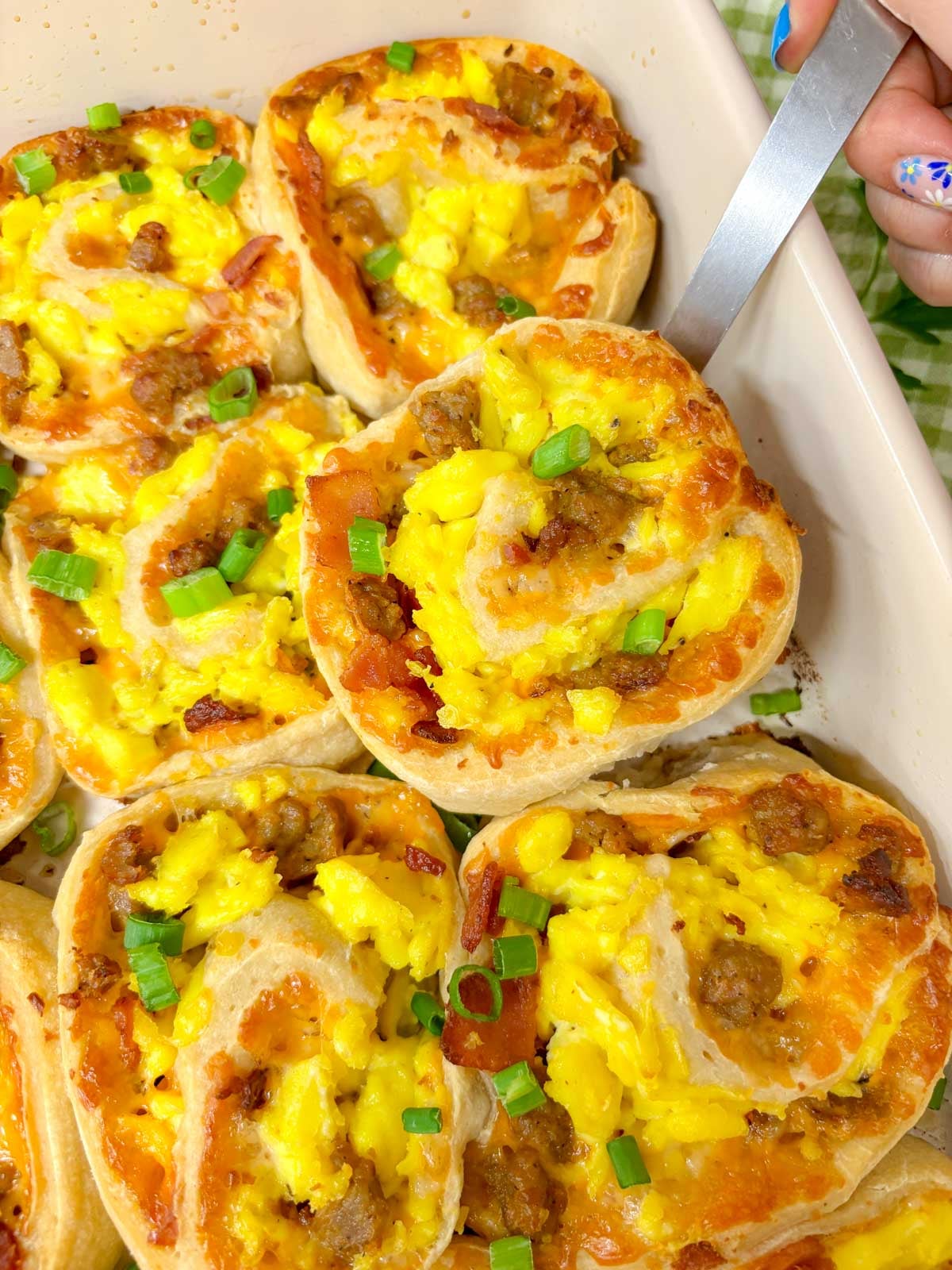 a sheet pan is filled with baked breakfast rolls that are garnished with green onion. one is held up with a spatula