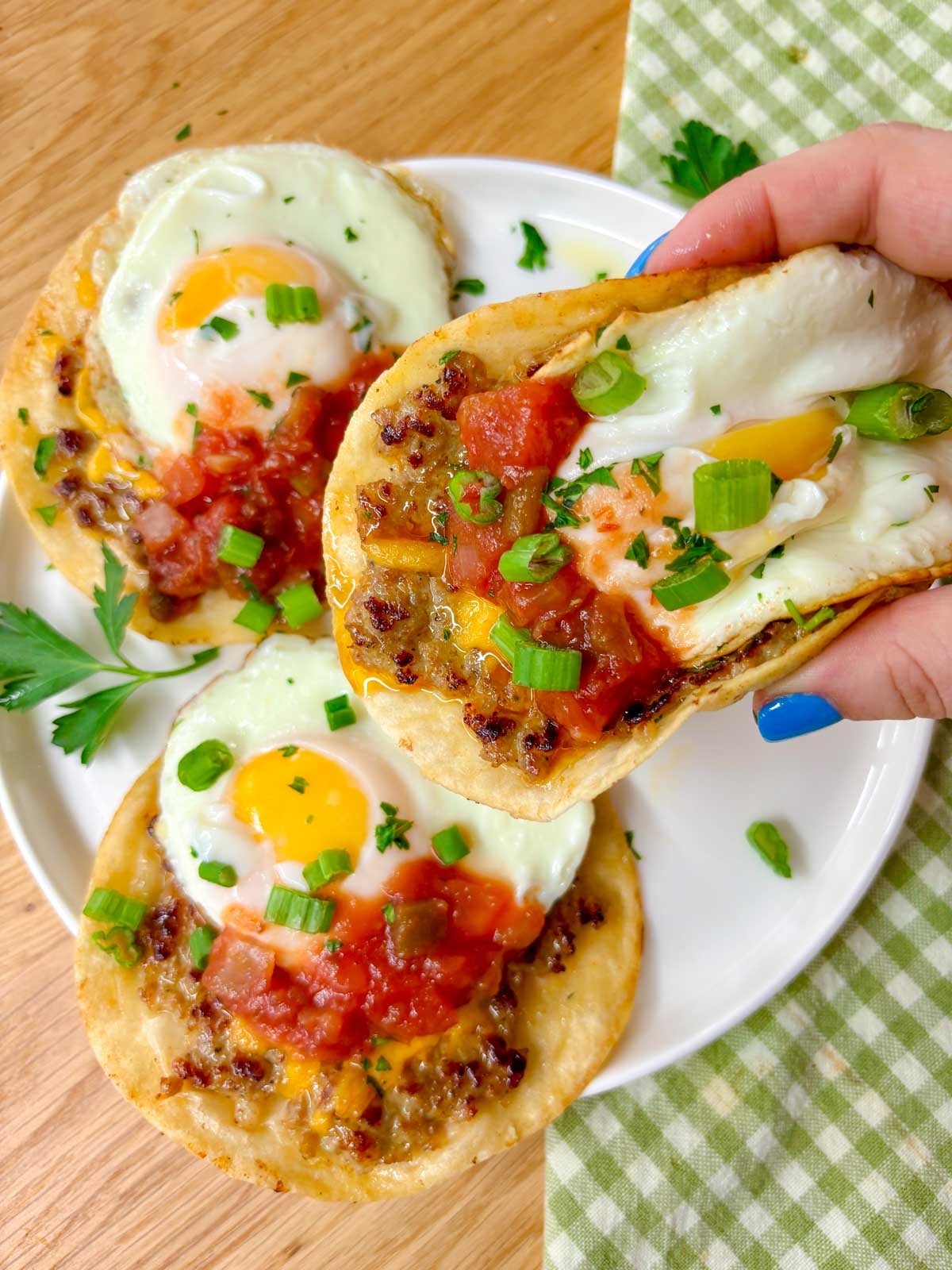 a white plate has three breakfast smash tacos on it garnished with salsa and green onions. one is held up close to the camera
